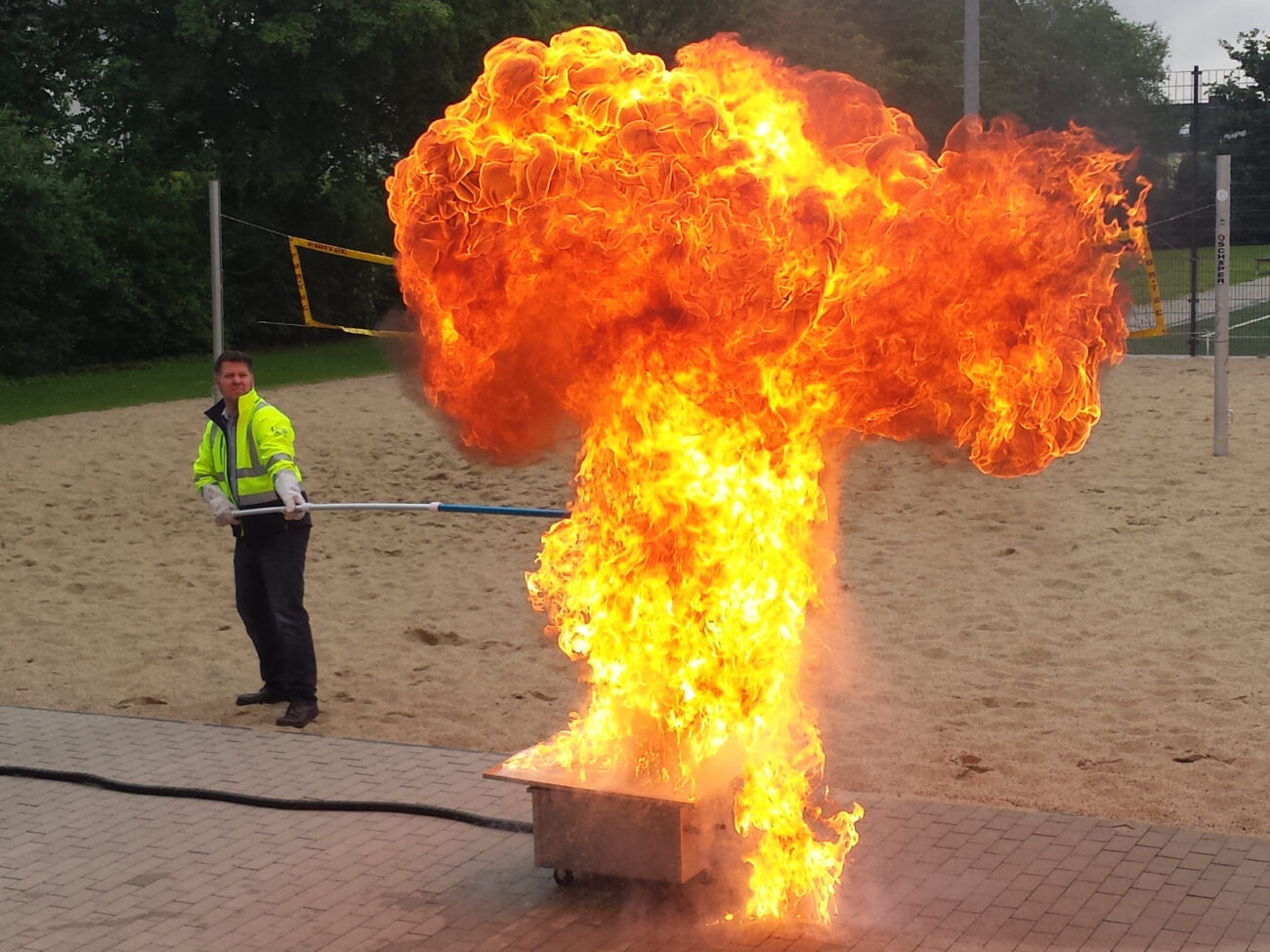 Ausbildung zum Brandschutzhelfer bei HSE-CAMPUS in Münster - Mann in gelber Warnjacke steht auf Beachvolleyballfeld und löscht große Stichflamme aus Brandvorrichtung